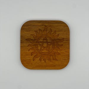 May include: A wooden coaster with a brown stain featuring a carved symbol of a five-pointed star inside a circle with flames around it.