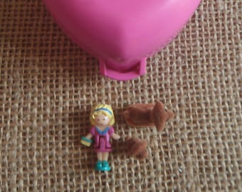 Può includere: Una custodia Polly Pocket rosa a forma di cuore con un'iscrizione dorata che recita "Polly Pocket". La custodia è aperta e all'interno ci sono tre piccole figure di plastica: una ragazza bionda con un vestito rosa e due cani marroni.