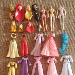 May include: A collection of disassembled doll parts, including heads, torsos, and various outfits. The outfits include dresses in pink, purple, and white, as well as pants and tops in yellow and red. The dolls' heads feature different hairstyles.