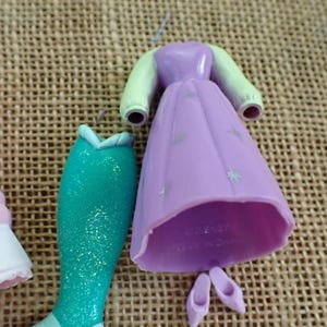 May include: A collection of doll clothing pieces, including a light purple dress with silver star accents, a teal mermaid tail, and pink shoes. The dress has light green sleeves. The items are made of plastic.