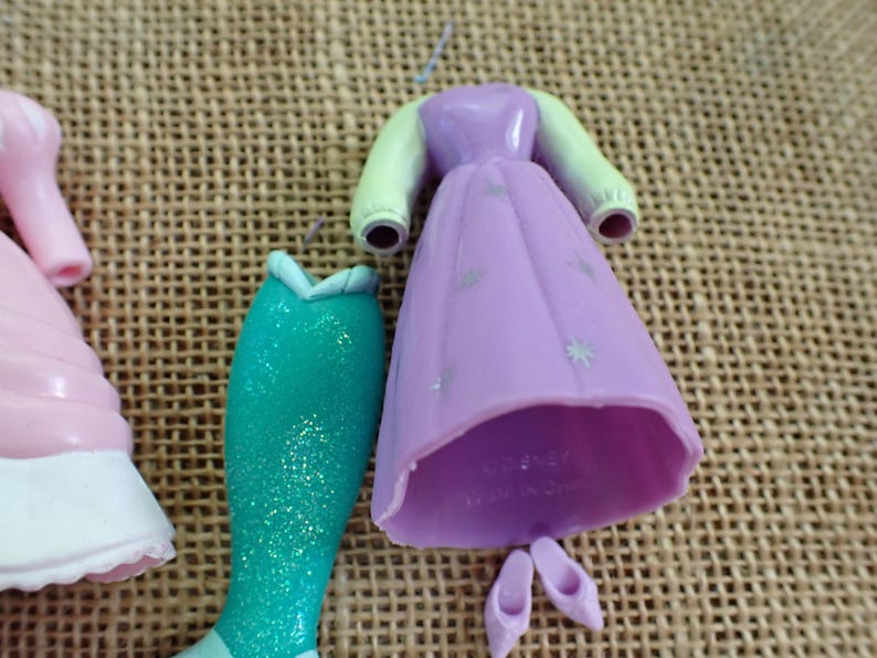 May include: A collection of doll clothing pieces, including a light purple dress with silver star accents, a teal mermaid tail, and pink shoes. The dress has light green sleeves. The items are made of plastic.
