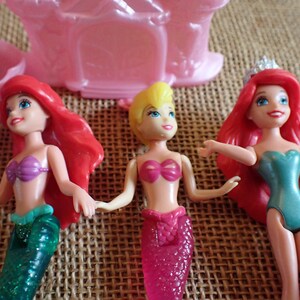May include: Three mermaid dolls with vibrant colors. One doll has red hair, a purple top, and a green tail. Another has blonde hair and a pink top and tail. The third doll has red hair, a teal top, and a pink tail. A pink castle is in the background.