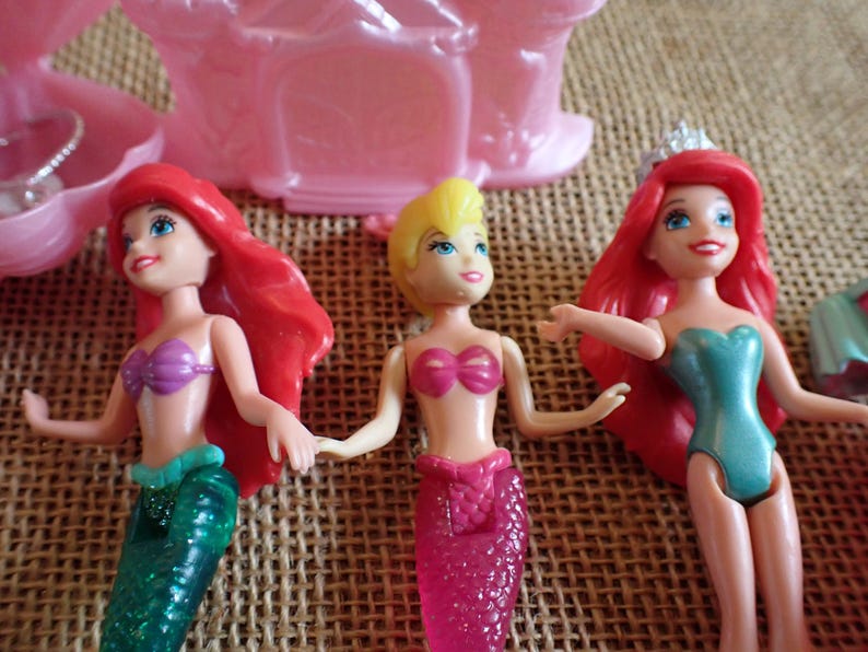 May include: Three mermaid dolls with vibrant colors. One doll has red hair, a purple top, and a green tail. Another has blonde hair and a pink top and tail. The third doll has red hair, a teal top, and a pink tail. A pink castle is in the background.