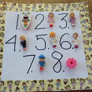 May include: A white paper with the numbers 1 through 8 written in black ink. Small plastic dolls and a toy boat are arranged next to the numbers. The paper is decorated with a border of cartoon girl illustrations.