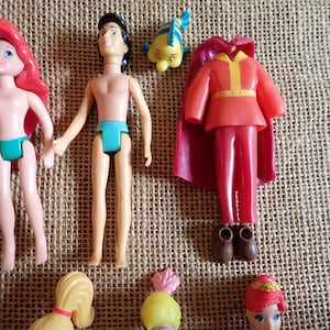 May include: A collection of miniature dolls and accessories, including a doll with red hair and a teal swimsuit, a doll with black hair and teal briefs, a red outfit with a cape, and a yellow fish figurine. The dolls are on a textured surface.