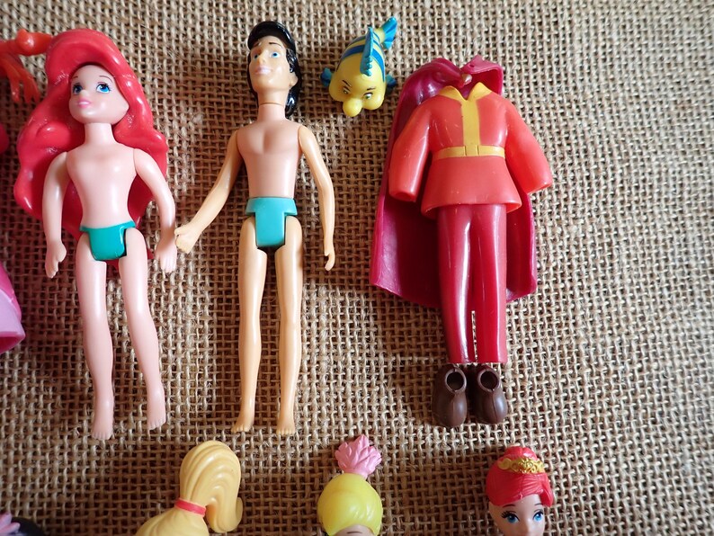 May include: A collection of miniature dolls and accessories, including a doll with red hair and a teal swimsuit, a doll with black hair and teal briefs, a red outfit with a cape, and a yellow fish figurine. The dolls are on a textured surface.