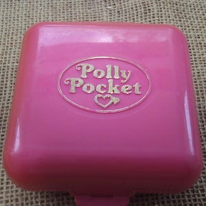 Op de afbeelding: Een felroze Polly Pocket compact, met een goudkleurig logo en hartontwerp. De vierkante compact heeft afgeronde randen en een kleine sluiting. Het ligt op een gestructureerd, neutraal gekleurd oppervlak.
