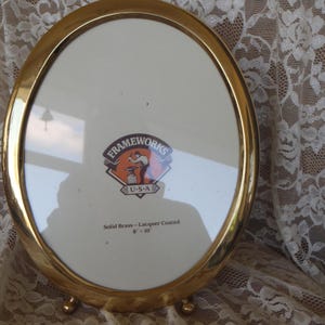 May include: Oval, gold-colored picture frame with a white interior. The frame has a brass finish and is supported by three small, round feet. The frame has the text "Frameworks U.S.A." and the dimensions 8 x 10 inches.