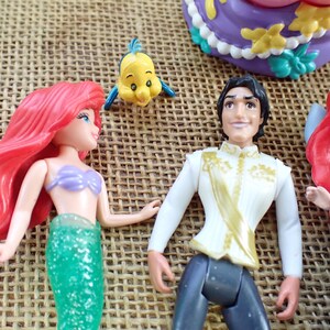 May include: A selection of Disney figurines featuring characters from The Little Mermaid. The figures include Ariel with red hair and a green tail, Prince Eric, and Flounder. A purple and yellow cake is also visible.