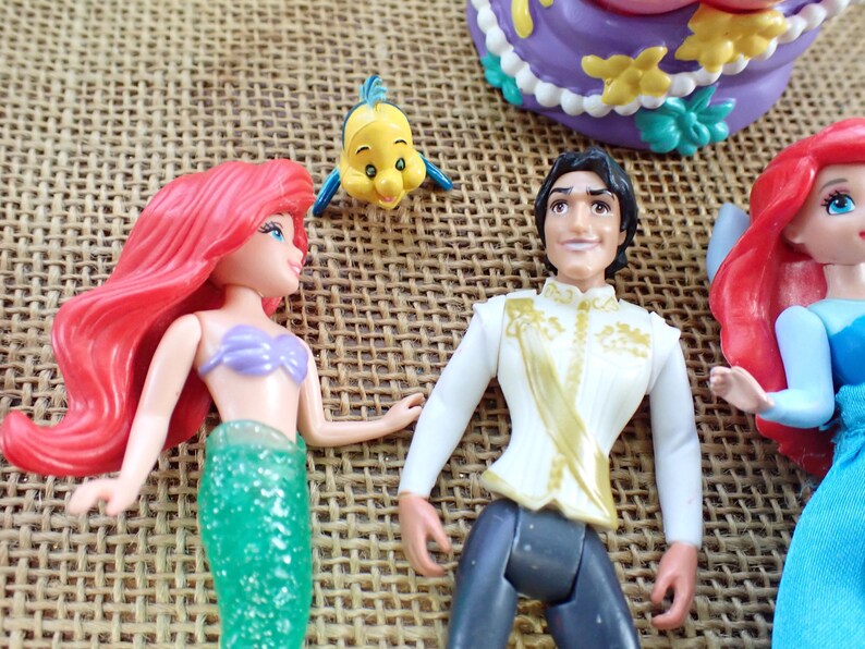 May include: A selection of Disney figurines featuring characters from The Little Mermaid. The figures include Ariel with red hair and a green tail, Prince Eric, and Flounder. A purple and yellow cake is also visible.