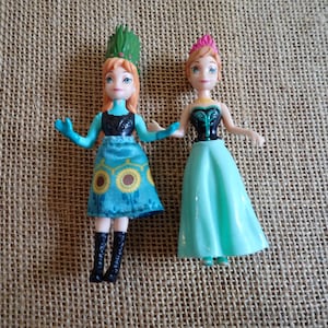 Peut inclure: Deux petites poup&eacute;es, chacune avec des cheveux roux et des yeux bleus, sont c&ocirc;te &agrave; c&ocirc;te. L'une porte une robe turquoise avec un motif de tournesol et un couvre-chef vert. L'autre porte une robe turquoise et un couvre-chef rose.