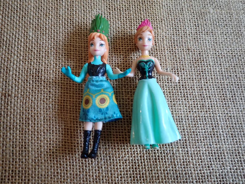 Peut inclure: Deux petites poup&eacute;es, chacune avec des cheveux roux et des yeux bleus, sont c&ocirc;te &agrave; c&ocirc;te. L'une porte une robe turquoise avec un motif de tournesol et un couvre-chef vert. L'autre porte une robe turquoise et un couvre-chef rose.