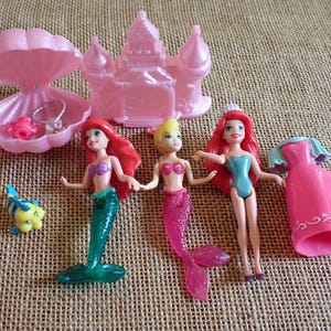 May include: A collection of pink and blue mermaid-themed toys. Includes three mermaid dolls with red, blonde, and red hair, a pink castle, a pink seashell with jewelry, a pink dress, and a yellow and blue fish.
