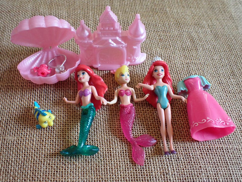 May include: A collection of pink and blue mermaid-themed toys. Includes three mermaid dolls with red, blonde, and red hair, a pink castle, a pink seashell with jewelry, a pink dress, and a yellow and blue fish.