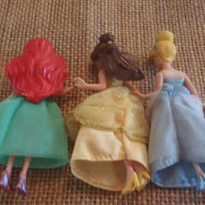 Op de afbeelding: Drie kleine poppen van Disney-prinsessen, van achteren gezien. Ariel heeft rood haar en een groene jurk. Belle heeft bruin haar en een gele jurk. Assepoester heeft blond haar en een blauwe jurk. Ze houden elkaars handen vast.