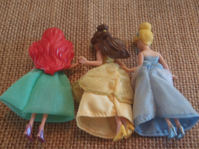 Op de afbeelding: Drie kleine poppen van Disney-prinsessen, van achteren gezien. Ariel heeft rood haar en een groene jurk. Belle heeft bruin haar en een gele jurk. Assepoester heeft blond haar en een blauwe jurk. Ze houden elkaars handen vast.