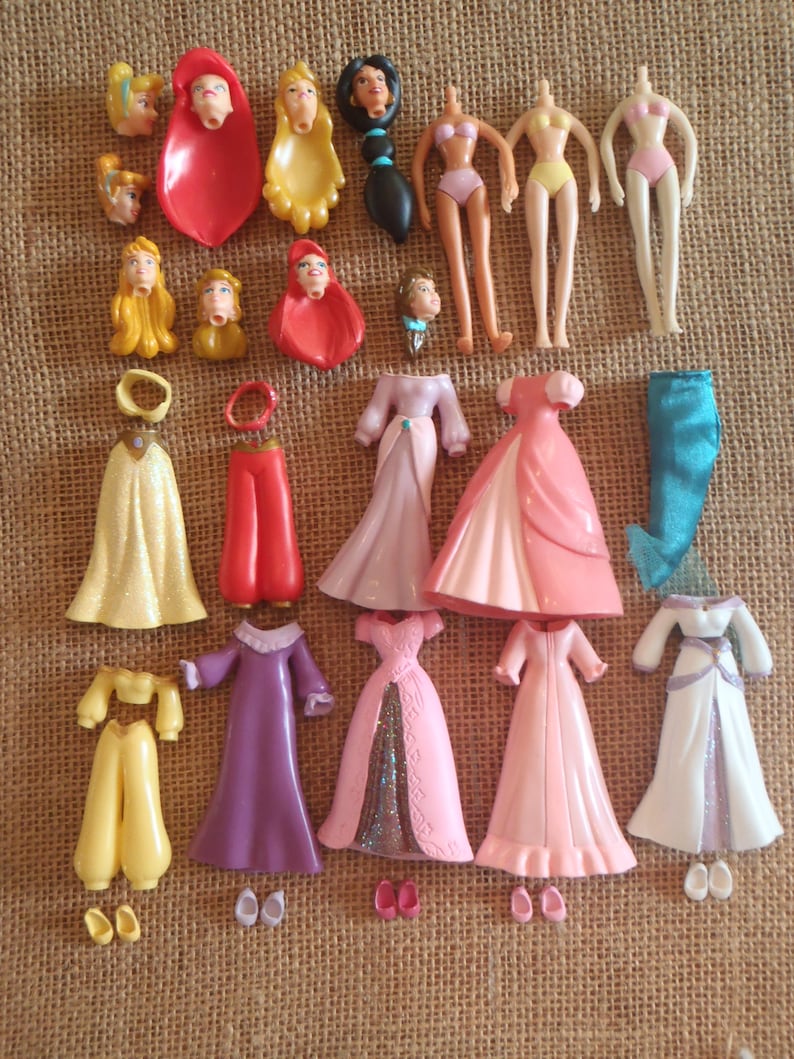 May include: A collection of disassembled doll parts, including heads, torsos, and various outfits. The outfits include dresses in pink, purple, and white, as well as pants and tops in yellow and red. The dolls' heads feature different hairstyles.