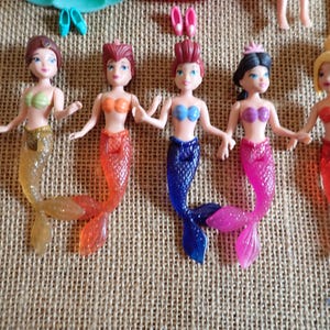 May include: Five mermaid dolls with colorful tails and tops. The dolls have different hair colors and styles, and are holding hands. Accessories like shoes and a tiara are visible.
