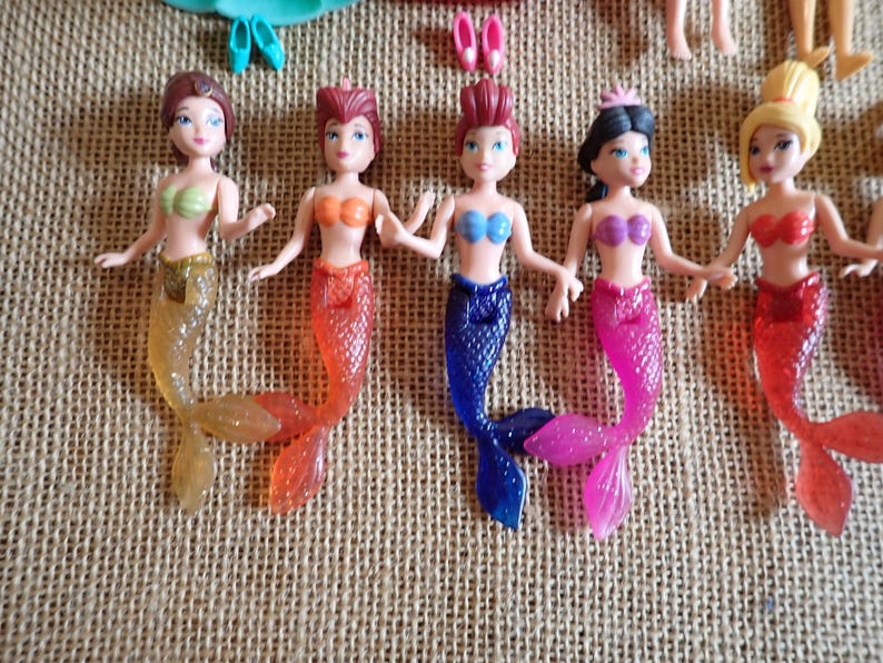 May include: Five mermaid dolls with colorful tails and tops. The dolls have different hair colors and styles, and are holding hands. Accessories like shoes and a tiara are visible.