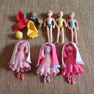 May include: A collection of doll accessories, including three doll bodies, three pink dresses with white capes, and various hair pieces in red, brown, and yellow. The dolls are small, with painted facial features.
