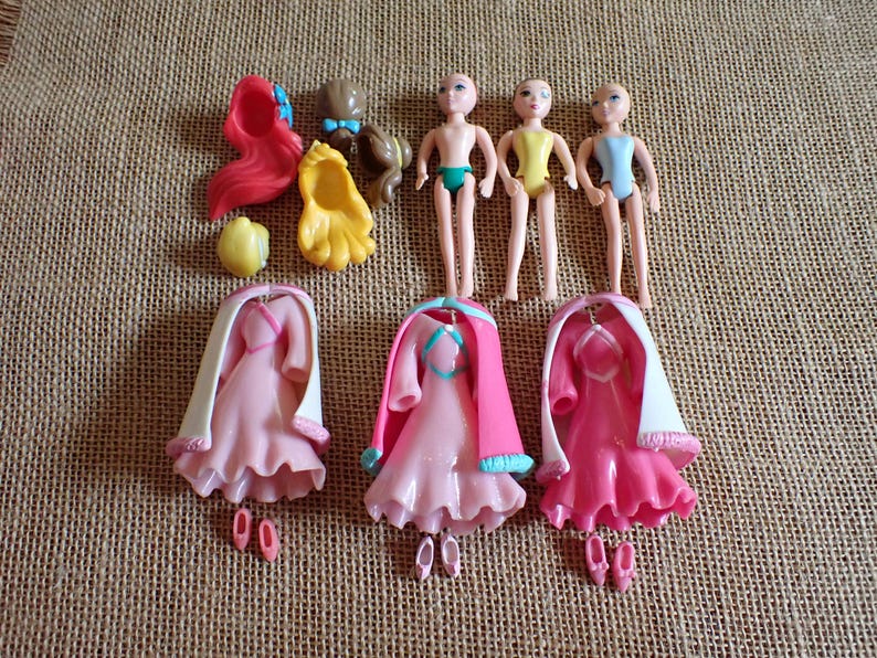 May include: A collection of doll accessories, including three doll bodies, three pink dresses with white capes, and various hair pieces in red, brown, and yellow. The dolls are small, with painted facial features.