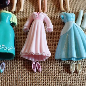 May include: A collection of miniature doll dresses and shoes in various colors. The dresses include a teal dress with white accents, a pink dress, and a light blue dress. Shoes are in matching colors.