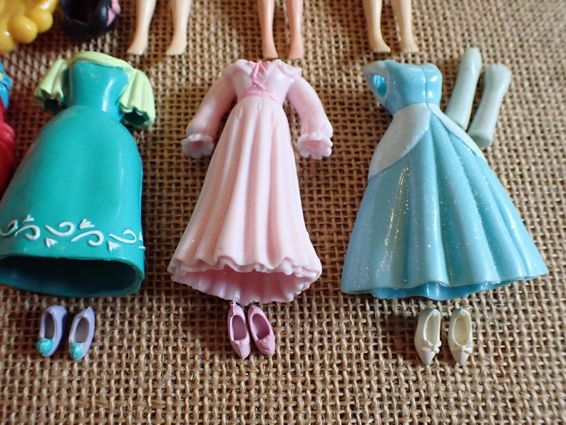 May include: A collection of miniature doll dresses and shoes in various colors. The dresses include a teal dress with white accents, a pink dress, and a light blue dress. Shoes are in matching colors.