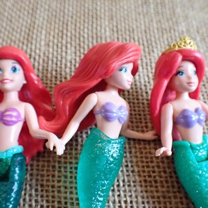 Puede incluir: Tres mu&ntilde;ecas de sirena con pelo rojo, ojos azules y tops de concha morada. Las mu&ntilde;ecas tienen colas de sirena verdes brillantes. Una mu&ntilde;eca lleva una corona dorada. Las mu&ntilde;ecas posan con los brazos extendidos.