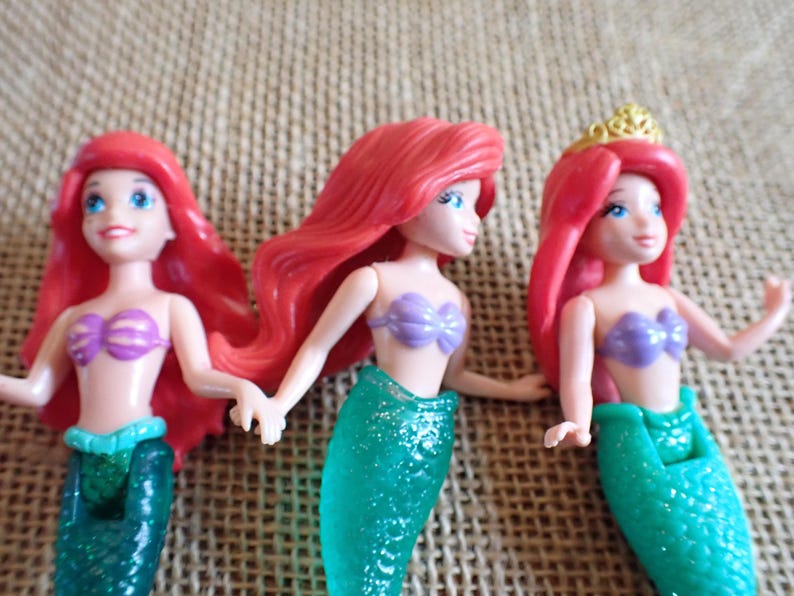 Puede incluir: Tres mu&ntilde;ecas de sirena con pelo rojo, ojos azules y tops de concha morada. Las mu&ntilde;ecas tienen colas de sirena verdes brillantes. Una mu&ntilde;eca lleva una corona dorada. Las mu&ntilde;ecas posan con los brazos extendidos.