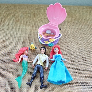 May include: A selection of Disney's The Little Mermaid toys, including Ariel, Prince Eric, and Flounder figurines. A pink clamshell case and Ariel in a blue dress are also featured. The toys are set against a neutral burlap background.