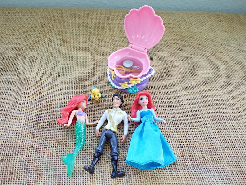 May include: A selection of Disney's The Little Mermaid toys, including Ariel, Prince Eric, and Flounder figurines. A pink clamshell case and Ariel in a blue dress are also featured. The toys are set against a neutral burlap background.