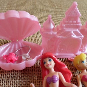 May include: A pink plastic playset featuring a seashell-shaped jewelry box with a necklace and a teapot, a castle, and two dolls with red and yellow hair. The toys are on a textured, tan surface.