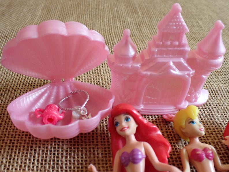 May include: A pink plastic playset featuring a seashell-shaped jewelry box with a necklace and a teapot, a castle, and two dolls with red and yellow hair. The toys are on a textured, tan surface.