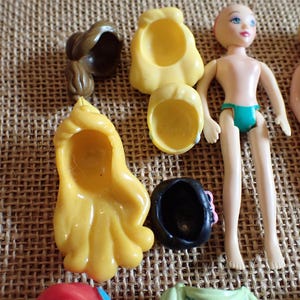 May include: A collection of doll parts, including two doll figures, several yellow and brown hair pieces, and a black hat with a pink bow. The dolls are nude, with one wearing teal underwear. The items are arranged on a textured, tan surface.