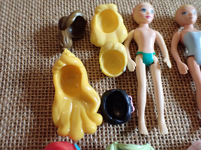 May include: A collection of doll parts, including two doll figures, several yellow and brown hair pieces, and a black hat with a pink bow. The dolls are nude, with one wearing teal underwear. The items are arranged on a textured, tan surface.