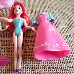 May include: Ariel and other mermaid dolls. Ariel has red hair, a blue swimsuit, and a tiara. A pink dress and a mermaid doll with a pink tail are also visible. The dolls are made of plastic and are on a textured surface.