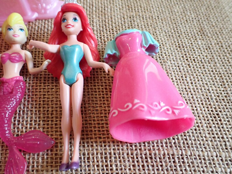 May include: Ariel and other mermaid dolls. Ariel has red hair, a blue swimsuit, and a tiara. A pink dress and a mermaid doll with a pink tail are also visible. The dolls are made of plastic and are on a textured surface.