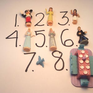 May include: A collection of small plastic figurines, including a Mickey Mouse figure, several princess figures, and a miniature blue bird. The figures are numbered 1 through 8 with a black marker on a white surface.