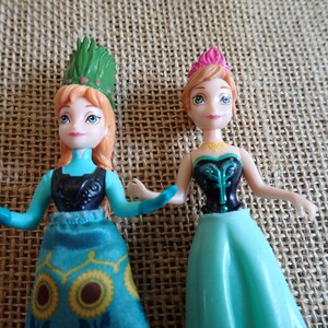Peut inclure: Deux petites poup&eacute;es, l'une avec une coiffe verte et l'autre avec une couronne rose. Les deux poup&eacute;es ont la peau claire, les yeux bleus et portent des robes dans les tons de bleu et de noir. Les poup&eacute;es sont pos&eacute;es sur un fond textur&eacute; et neutre.