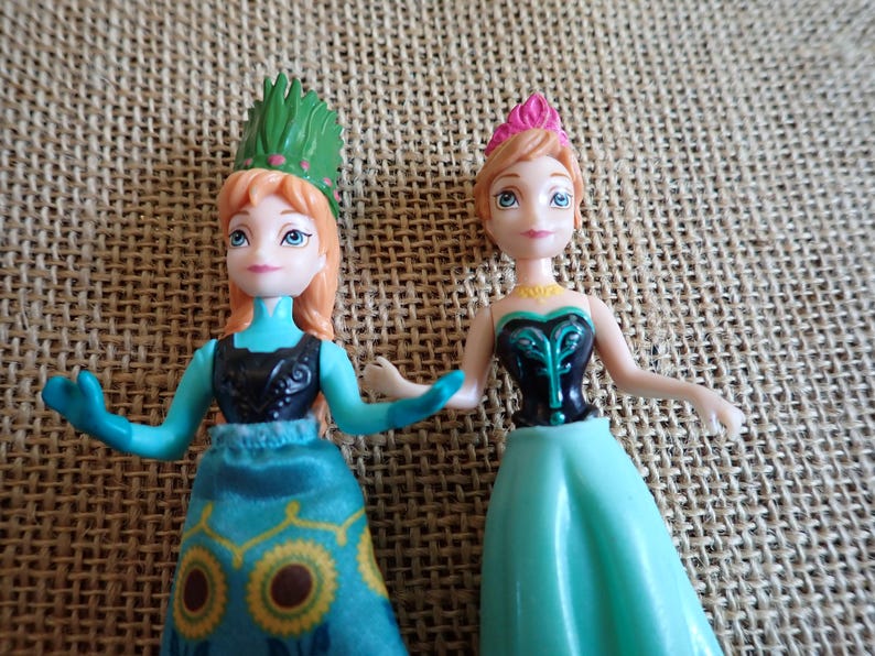 Peut inclure: Deux petites poup&eacute;es, l'une avec une coiffe verte et l'autre avec une couronne rose. Les deux poup&eacute;es ont la peau claire, les yeux bleus et portent des robes dans les tons de bleu et de noir. Les poup&eacute;es sont pos&eacute;es sur un fond textur&eacute; et neutre.