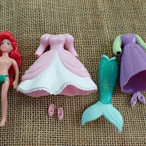 May include: A doll with red hair, green underwear, and various outfits. Includes a pink dress, a purple dress, a teal mermaid tail, and pink shoes. The doll is a classic toy.
