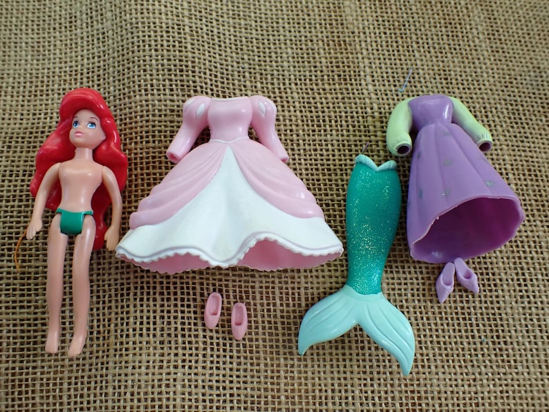 May include: A doll with red hair, green underwear, and various outfits. Includes a pink dress, a purple dress, a teal mermaid tail, and pink shoes. The doll is a classic toy.