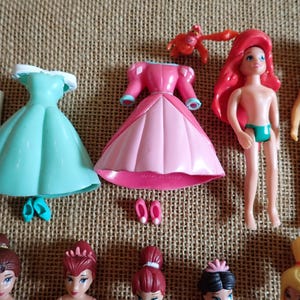 May include: A collection of miniature plastic dolls and accessories. Includes a teal dress, pink dress, Ariel doll, Prince doll, and other doll figures. Accessories include shoes and a crab figurine.