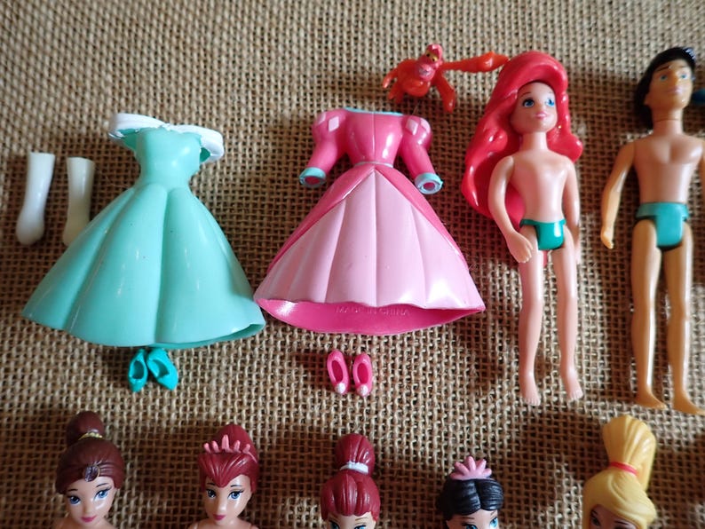 May include: A collection of miniature plastic dolls and accessories. Includes a teal dress, pink dress, Ariel doll, Prince doll, and other doll figures. Accessories include shoes and a crab figurine.