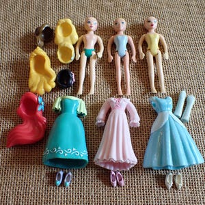 May include: A collection of miniature doll parts, including three doll figures, three dresses in teal, pink, and blue, and various accessories like shoes and wigs. The dolls are nude with painted features.