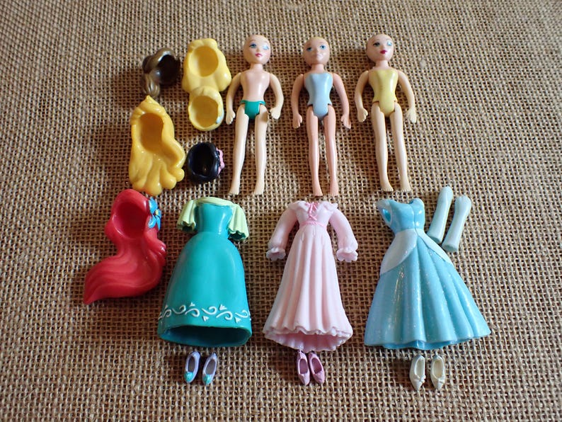 May include: A collection of miniature doll parts, including three doll figures, three dresses in teal, pink, and blue, and various accessories like shoes and wigs. The dolls are nude with painted features.