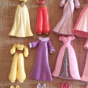 May include: A collection of miniature doll clothing and accessories in various colors. Includes yellow, red, pink, and purple dresses, pants, tops, and shoes. The items are arranged on a textured, tan surface.