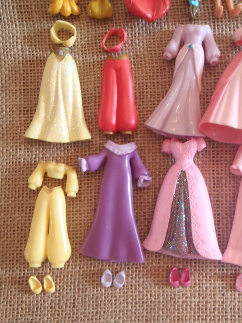 May include: A collection of miniature doll clothing and accessories in various colors. Includes yellow, red, pink, and purple dresses, pants, tops, and shoes. The items are arranged on a textured, tan surface.