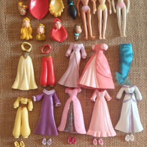 May include: A collection of vintage doll parts, including heads, torsos, and various outfits. The outfits include dresses in shades of pink, white, and purple, as well as pants and tops in yellow and red. The dolls are missing their heads.