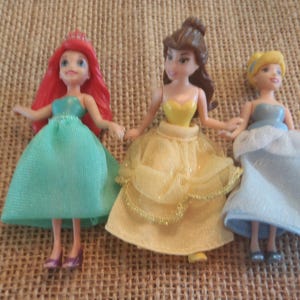 Op de afbeelding: Drie miniatuur prinsessenpoppen. Ariel draagt een zeegroene jurk en heeft rood haar. Belle draagt een gele jurk. Assepoester draagt een lichtblauwe jurk. Alle drie de poppen houden elkaars hand vast en staan samen.